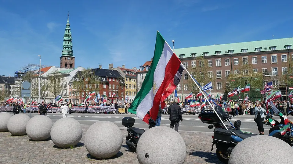 Marchers with Iranian lion and sun flags cross a cobbled square in Copenhagen under a blue sky