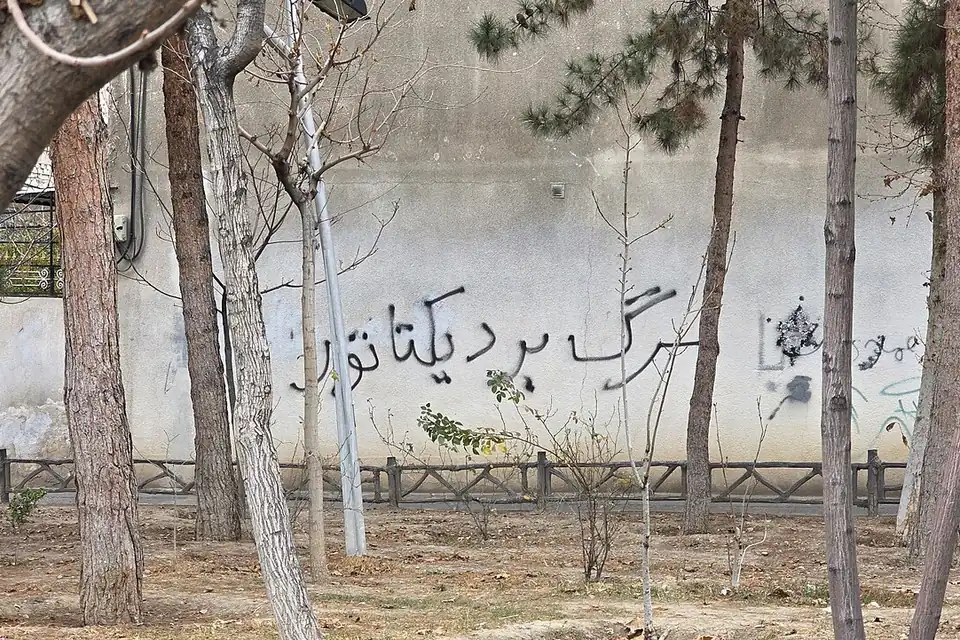 Persian graffiti reading Death to the dictator sprayed on a pale wall behind a row of trees