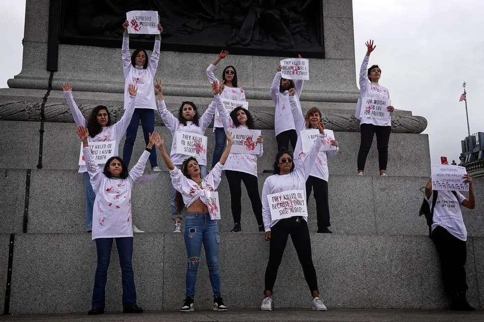 Women in white shirts marked with red handprints stand on a monument plinth holding placards