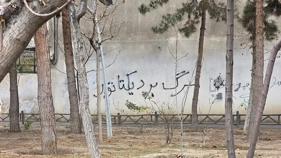 Persian graffiti reading Death to the dictator sprayed on a pale wall behind a row of trees
