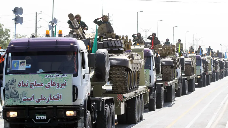A column of trucks carrying tanks with IRGC personnel aboard drives down a city avenue