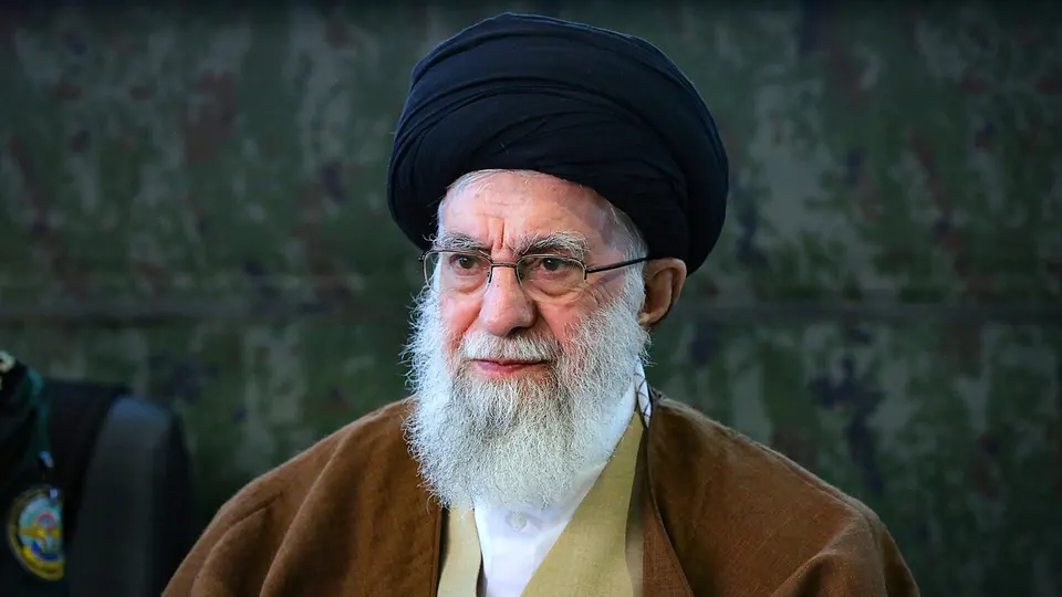 Ali Khamenei, in a black turban, brown cloak and glasses, seated in front of a camouflage backdrop