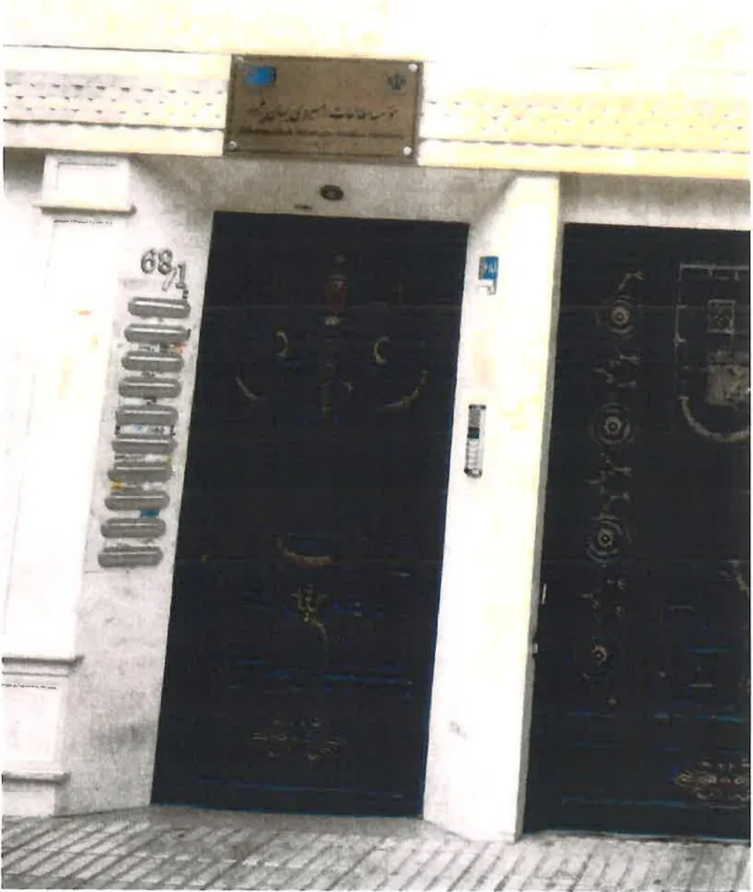 Two dark metal doors under a nameplate in Farsi, beside a column of door buzzers
